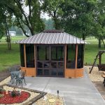 Hexagonal gazebo on concrete with trees, a bench swing, and a garden table set in a yard.