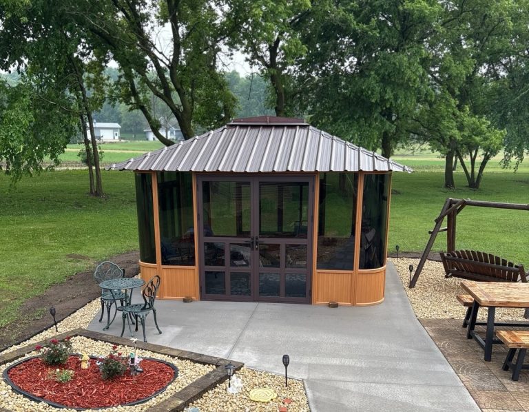 Hexagonal gazebo on concrete with trees, a bench swing, and a garden table set in a yard.