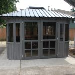 Gray backyard gazebo with glass windows and a metal roof on a concrete patio.
