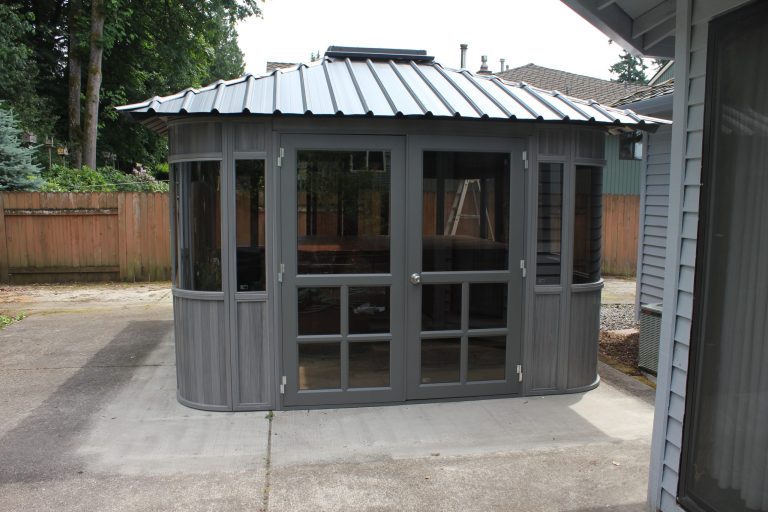 Gray backyard gazebo with glass windows and a metal roof on a concrete patio.