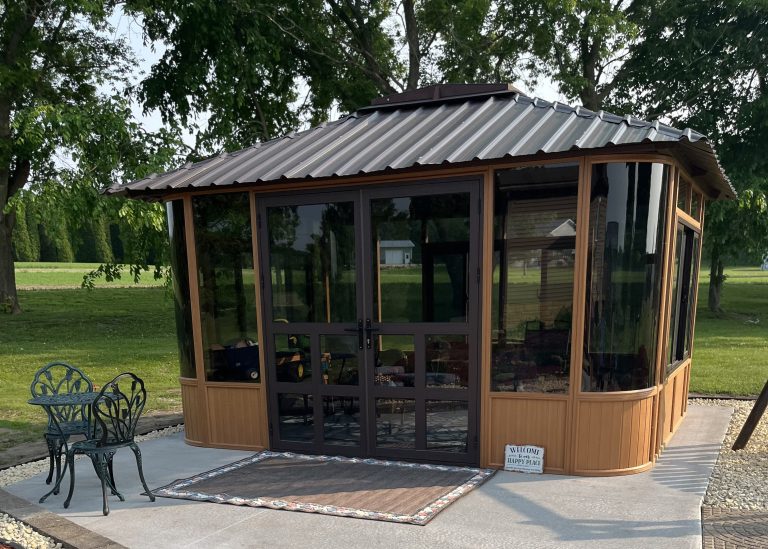 Brown gazebo with black framed glass doors and windows, metal roof, patio furniture, surrounded by trees.