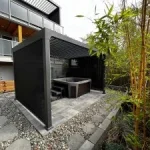 Modern outdoor patio with pergola, hot tub, stone path, and bamboo plants in a backyard setting.