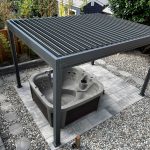 Outdoor hot tub under a metal pergola, surrounded by gravel, tiles, and a wooden fence.