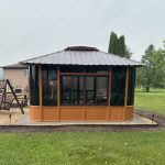 Wooden gazebo with dark windows and metal roof in a garden setting with grass and trees.