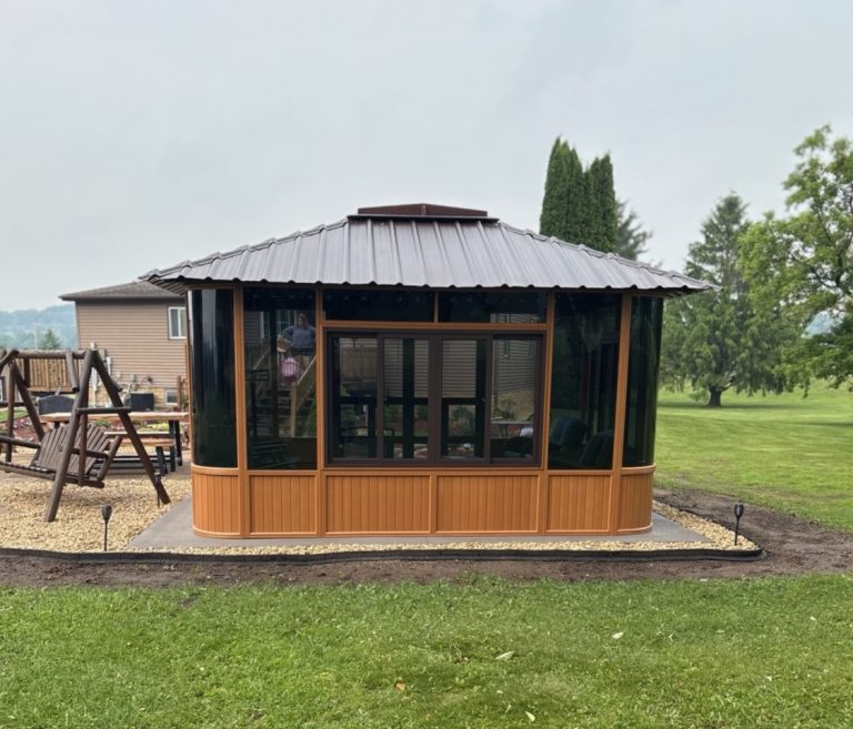 Wooden gazebo with dark windows and metal roof in a garden setting with grass and trees.