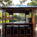A wooden gazebo bar setup with stools and a covered grill, surrounded by a garden with a wooden fence.