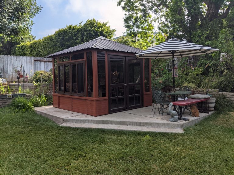 Garden with a gazebo, striped umbrella, metal chairs, and a red bench on a concrete patio surrounded by greenery.