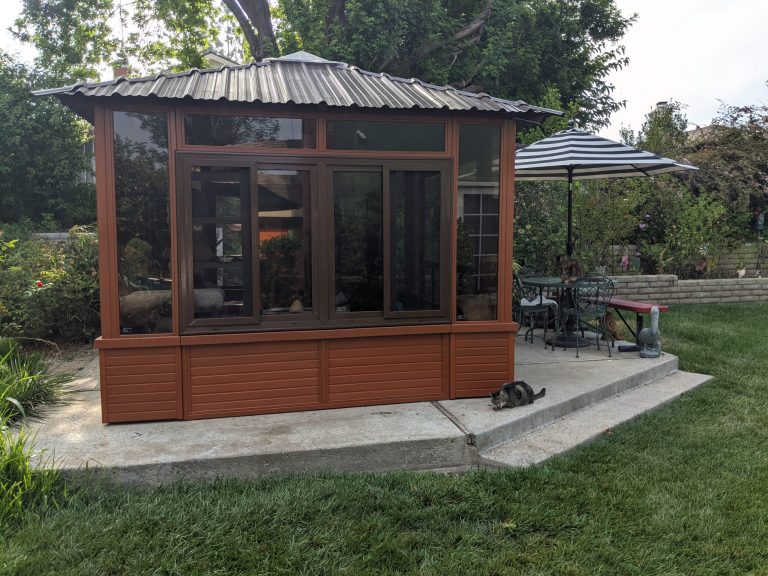 Small wooden gazebo with windows, a striped umbrella, patio furniture, and two cats on the concrete patio.