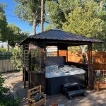 Gazebo with a hot tub inside, surrounded by trees and a fence, on a sunny day.
