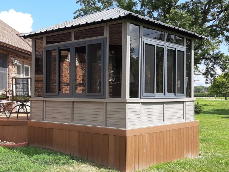 A small sunroom with large windows and a metal roof attached to a brick house, surrounded by grass and trees.