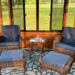Wicker chairs with blue cushions and footrests in a sunroom with large windows overlooking a green yard.