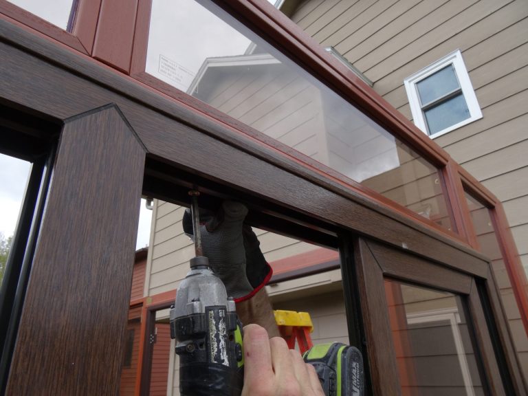 Person using a power drill to install screws on a wooden frame with a house in the background.