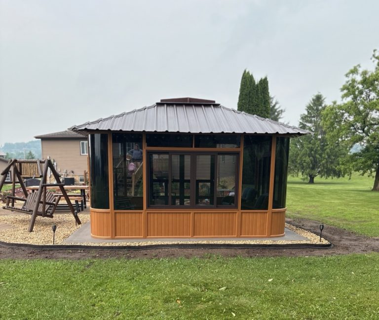 A wooden gazebo with glass windows, set in a garden with a wooden swing and trees in the background.