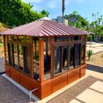 A wooden gazebo with glass windows, situated in a landscaped garden with plants and a tiled walkway.
