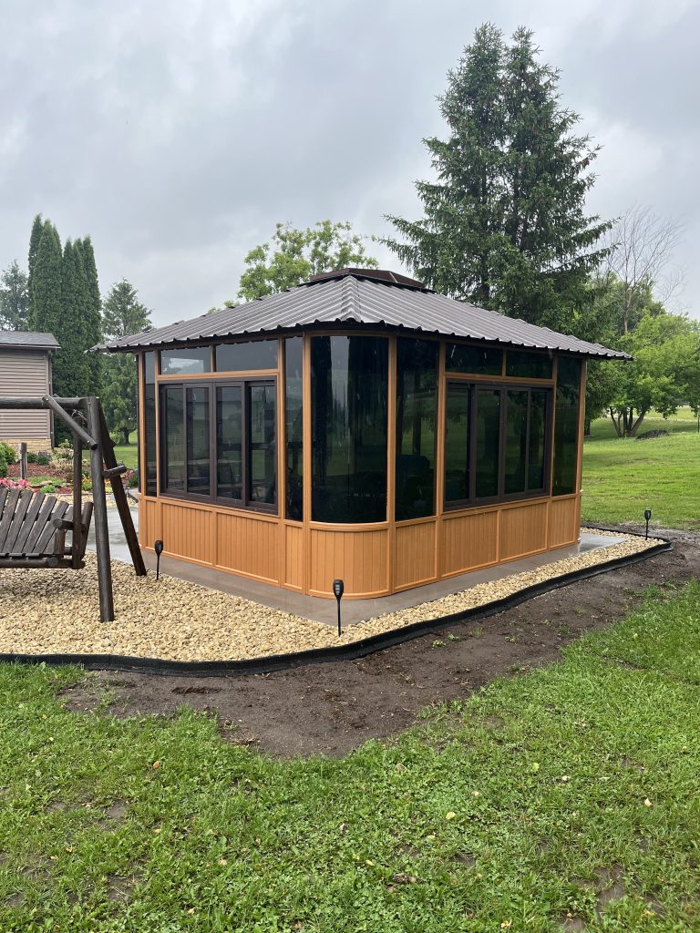 A wooden gazebo with a black roof and glass windows surrounded by a gravel area and grass in a garden.