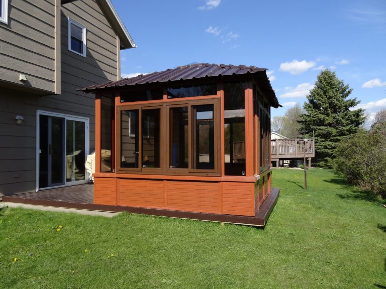 Brown gazebo with windows on a wooden deck, attached to a house with a green lawn and blue sky.