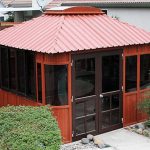 The Aspen 12x14, a wooden gazebo with a red roof, large windows, and double glass doors, is on sale. It sits in a backyard surrounded by bushes, rocks, and has a paved pathway leading to its entrance.