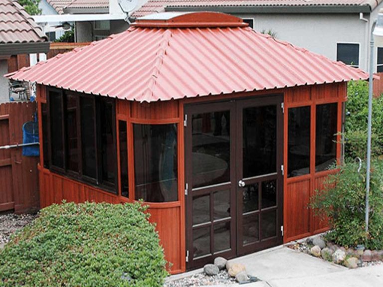The Aspen 12x14, a wooden gazebo with a red roof, large windows, and double glass doors, is on sale. It sits in a backyard surrounded by bushes, rocks, and has a paved pathway leading to its entrance.