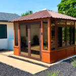 Small wooden gazebo with glass windows and a metal roof beside a house, surrounded by black stone landscaping.