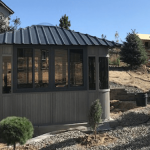 Small gray gazebo with glass windows, metal roof, surrounded by a rocky landscape and small trees.