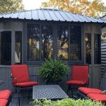 Gazebo with red-cushioned chairs and a black table on a deck, surrounded by greenery.