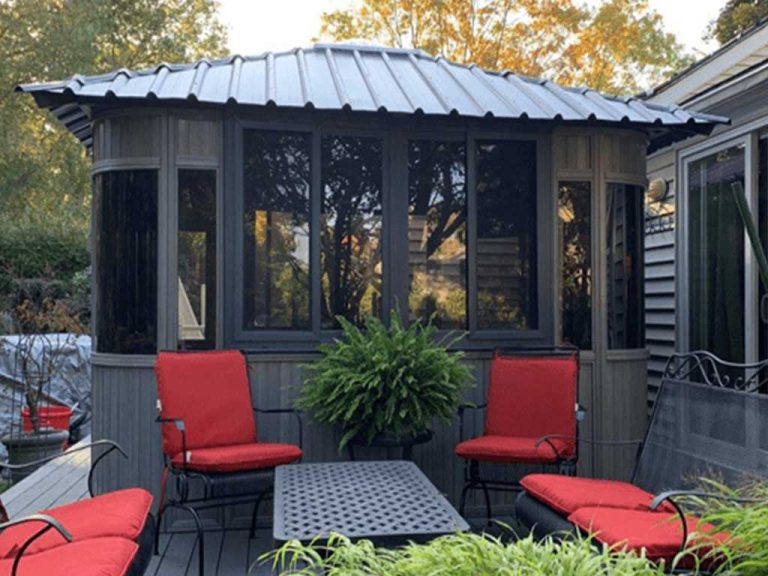 Gazebo with red-cushioned chairs and a black table on a deck, surrounded by greenery.