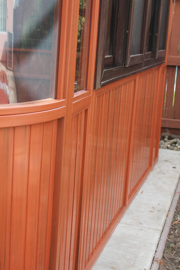 Close Up Of An Orange Wooden Exterior Wall With Windows And A Concrete Path Alongside.