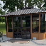 Brown gazebo with black framed glass doors and windows, metal roof, patio furniture, surrounded by trees.