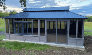 Gray, Modern Gazebo With Large Windows And A Blue Metal Roof Sits On A Concrete Slab In A Grassy Area.