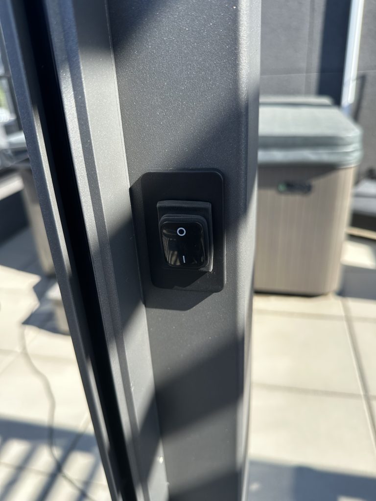 Close-up of a black rocker switch on an outdoor metal post, with tiled flooring and shadows visible.