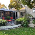 Backyard with a patio, red-cushioned chairs, plants in raised beds, and a gazebo beside a house.
