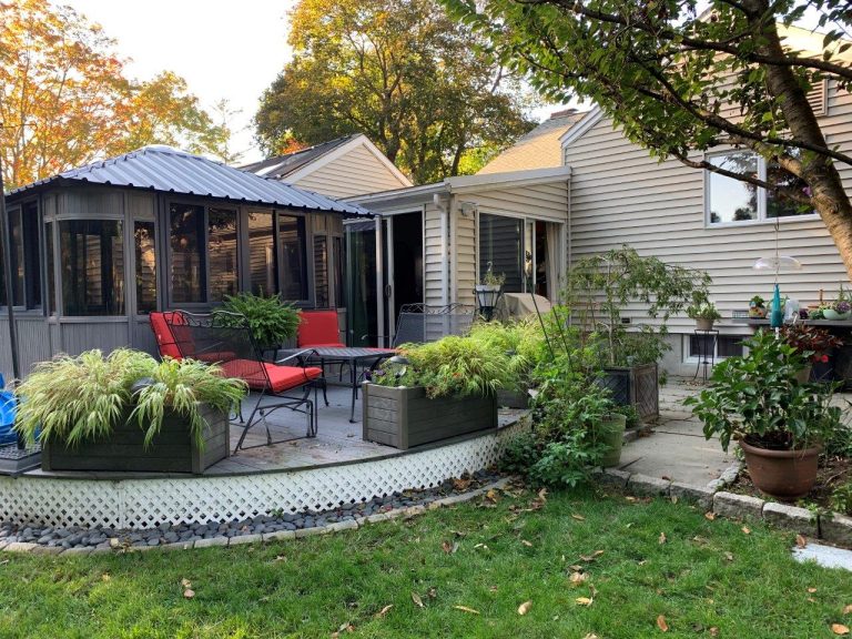 Backyard with a patio, red-cushioned chairs, plants in raised beds, and a gazebo beside a house.