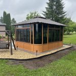 A wooden gazebo with a black roof and glass windows surrounded by a gravel area and grass in a garden.