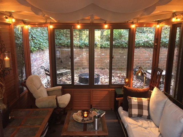 Cozy sunroom with string lights, armchairs, a sofa, and a coffee table, overlooking a leafy brick-walled outdoor area.