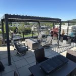 Rooftop patio with chairs, a pergola, planters, and a view of the cityscape and trees in bright sunlight.