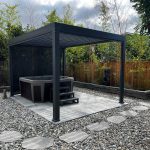 Outdoor hot tub under a modern pergola, surrounded by pebbles and stepping stones, bordered by bamboo and wooden fencing.