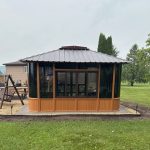 A wooden gazebo with glass windows, set in a garden with a wooden swing and trees in the background.
