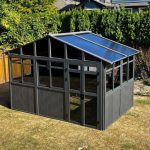 Small gray greenhouse with transparent roof panels in a backyard with a wooden fence and greenery.