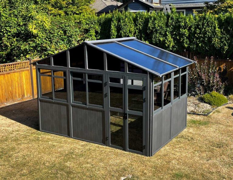 Small gray greenhouse with transparent roof panels in a backyard with a wooden fence and greenery.