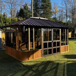 Wooden gazebo with glass windows and metal roof on a sunny lawn, near trees and a small shed.
