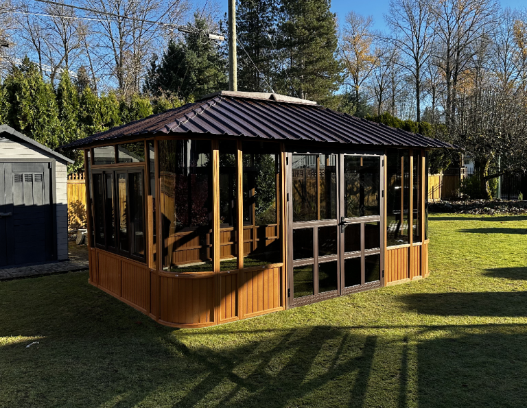 Wooden gazebo with glass windows and metal roof on a sunny lawn, near trees and a small shed.