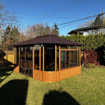 Wooden gazebo with glass windows on a sunny lawn, surrounded by a wooden fence and trees with a clear blue sky.