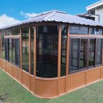 Wooden gazebo with large windows on green grass, under a blue sky.