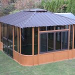 A brown gazebo with glass windows on a grassy lawn, surrounded by a wooden fence and greenery.