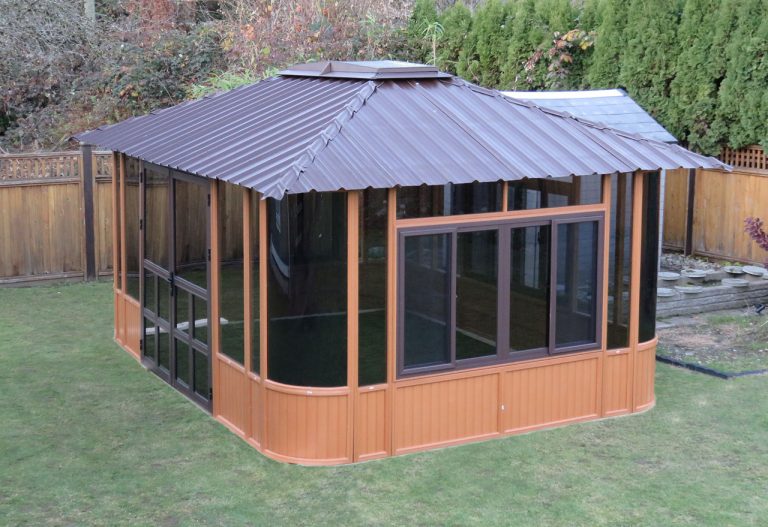 A brown gazebo with glass windows on a grassy lawn, surrounded by a wooden fence and greenery.