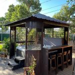 A wooden gazebo with a metal roof, enclosing a hot tub and bar seating, set in a lush, green backyard.