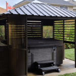 Wooden gazebo with metal roof, housing a hot tub, located in a landscaped backyard with grass and bushes.