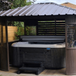 Wooden gazebo with a metal roof housing a hot tub, surrounded by a wooden fence and plants.