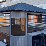 Covered hot tub with a louvered gazebo, bar counter, and stools on a patio with nearby house background.