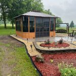 Wooden gazebo with glass panels on a rainy day, surrounded by trees, plants, and a landscaped garden path.